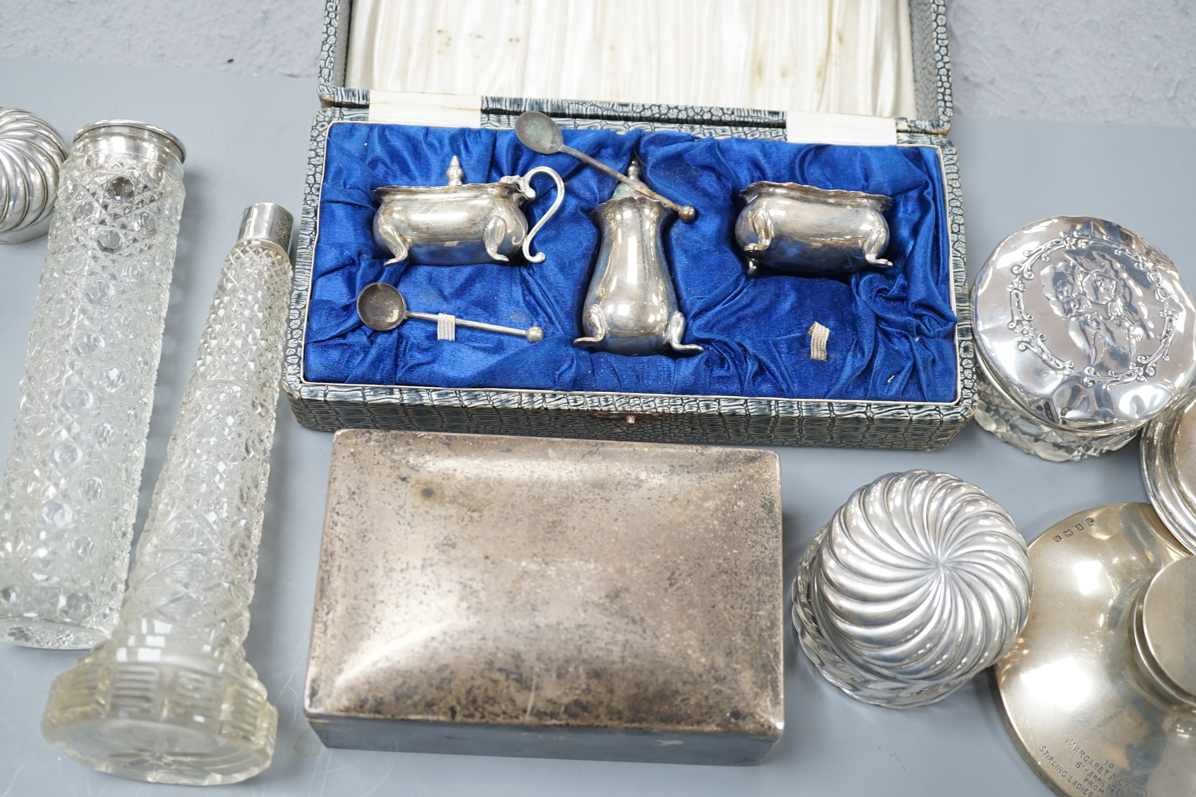 Assorted silver items including a cigarette box, a cased silver condiment set, two capstan inkwells, a pair of Edwardian dwarf candlesticks and four silver mounted glass toilet jars.